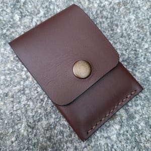 May include: A dark brown leather card holder featuring a rectangular flap secured by a single circular brass-toned metal snap button. The edges of the pouch are joined with visible stitching along the side and bottom, creating a compact pocket.