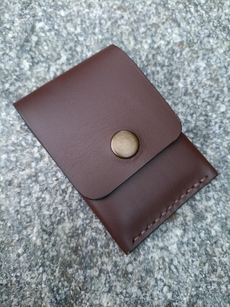May include: A dark brown leather card holder featuring a rectangular flap secured by a single circular brass-toned metal snap button. The edges of the pouch are joined with visible stitching along the side and bottom, creating a compact pocket.