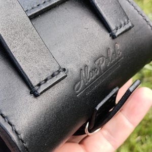May include: Close-up of a black leather case with straps and stitching. The embossed text "Alex Pelak" is visible on the leather. The case has a silver metal ring and is held in a hand.