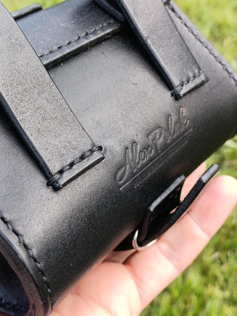May include: Close-up of a black leather case with straps and stitching. The embossed text "Alex Pelak" is visible on the leather. The case has a silver metal ring and is held in a hand.