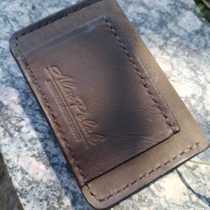 May include: A rectangular dark brown leather card holder featuring visible stitching along the edges and a debossed logo that reads Alex Polak Handmade Leather Goods. The leather has a smooth texture with a slightly darker central panel.