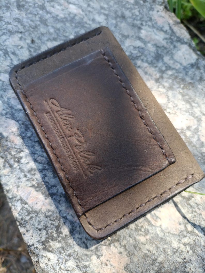 May include: A rectangular dark brown leather card holder featuring visible stitching along the edges and a debossed logo that reads Alex Polak Handmade Leather Goods. The leather has a smooth texture with a slightly darker central panel.