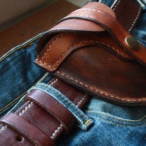 K&ouml;nnte beinhalten: Eine braune Lederholster mit geschwungenem Design, mit sichtbaren N&auml;hten und einem Messingknopfverschluss. Die Holster ist &uuml;ber einem braunen Lederg&uuml;rtel und einer blauen Jeans positioniert und zeigt eine rustikale &Auml;sthetik.