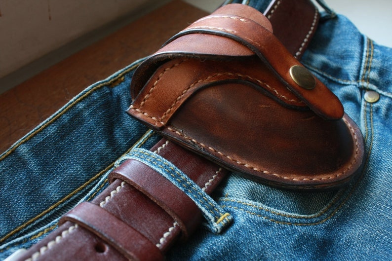 K&ouml;nnte beinhalten: Eine braune Lederholster mit geschwungenem Design, mit sichtbaren N&auml;hten und einem Messingknopfverschluss. Die Holster ist &uuml;ber einem braunen Lederg&uuml;rtel und einer blauen Jeans positioniert und zeigt eine rustikale &Auml;sthetik.