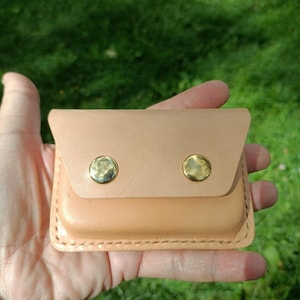 May include: A rectangular light tan leather pouch featuring a fold-over flap secured by two polished gold-tone metal snap buttons. The edges of the pouch are reinforced with visible thick stitching along the perimeter of the leather construction.