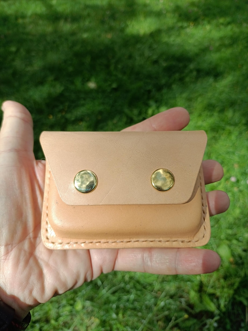 May include: A rectangular light tan leather pouch featuring a fold-over flap secured by two polished gold-tone metal snap buttons. The edges of the pouch are reinforced with visible thick stitching along the perimeter of the leather construction.