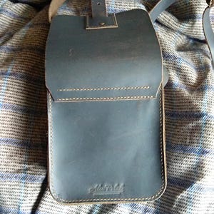 Pu&ograve; includere: Una borsa a tracolla in pelle blu scuro con una tracolla in pelle marrone e una fibbia. La borsa ha una patta con un bordo cucito e una piccola tasca sul davanti. La borsa &egrave; etichettata "Ale-Polk" in lettere dorate.