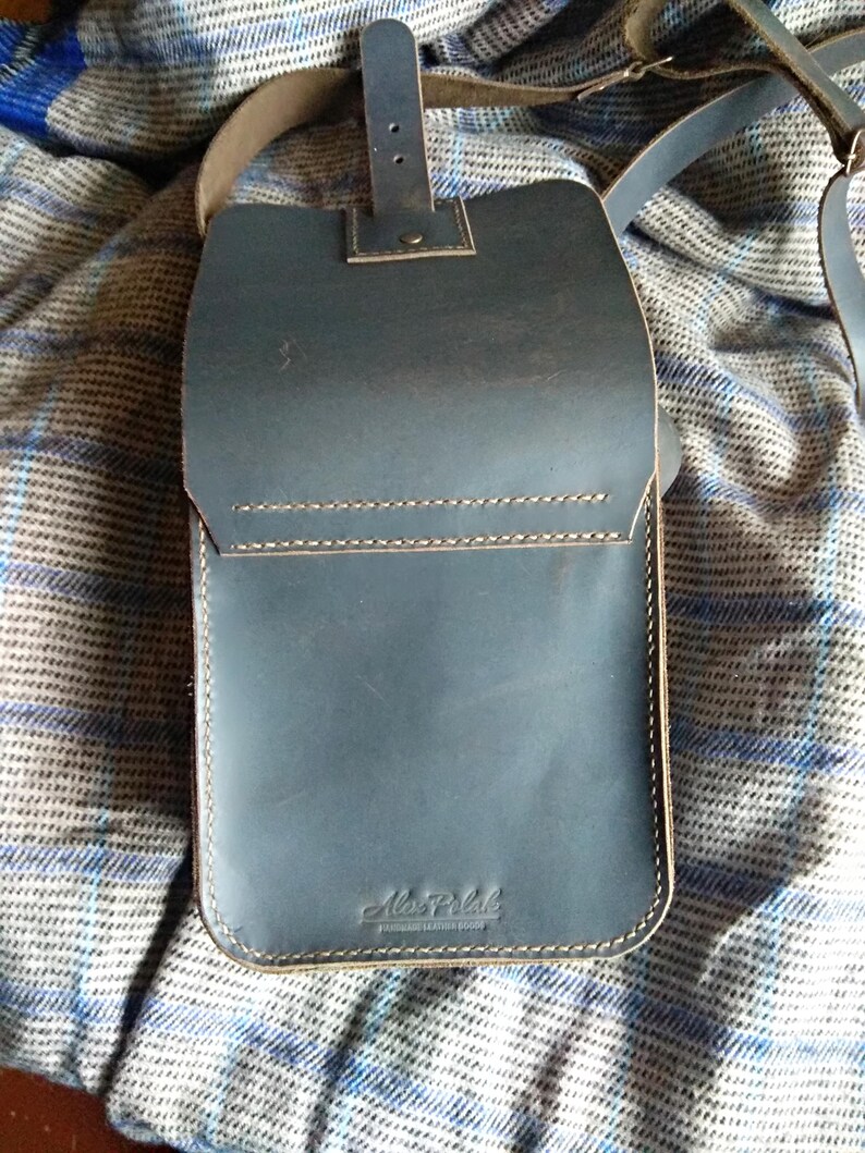 Pu&ograve; includere: Una borsa a tracolla in pelle blu scuro con una tracolla in pelle marrone e una fibbia. La borsa ha una patta con un bordo cucito e una piccola tasca sul davanti. La borsa &egrave; etichettata "Ale-Polk" in lettere dorate.
