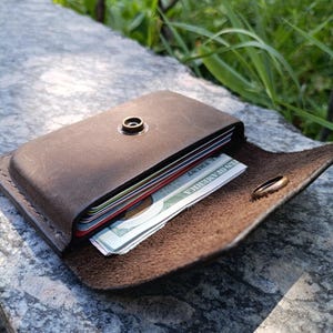 May include: A dark brown leather card holder with a brass snap button closure. The wallet is partially open, revealing a stack of credit cards and a United States one hundred dollar bill with the text United States of America visible.