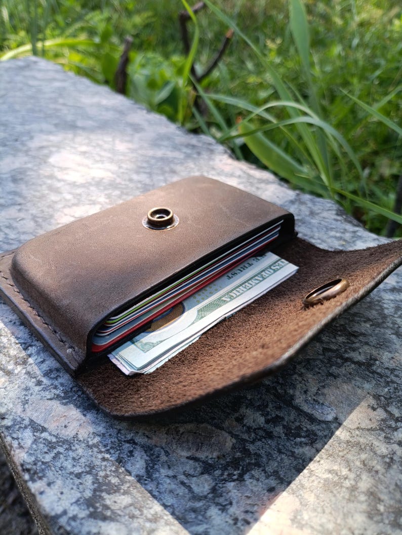 May include: A dark brown leather card holder with a brass snap button closure. The wallet is partially open, revealing a stack of credit cards and a United States one hundred dollar bill with the text United States of America visible.
