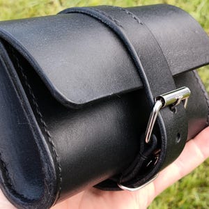 May include: A black leather pouch with a flap closure and a buckle strap. The pouch is cylindrical and appears to be hand-stitched. The strap is secured with a silver-colored metal buckle.