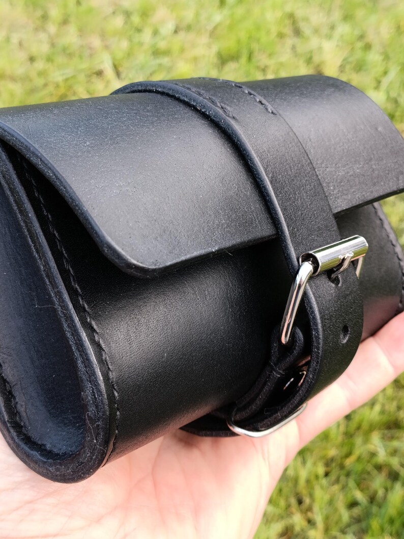 May include: A black leather pouch with a flap closure and a buckle strap. The pouch is cylindrical and appears to be hand-stitched. The strap is secured with a silver-colored metal buckle.