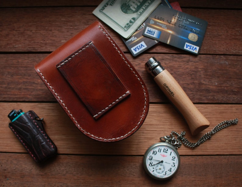 May include: A brown leather pouch with a white stitch detail, a pocket knife with a wooden handle, a pocket watch with a silver chain, and a lighter with a black and blue patterned design.