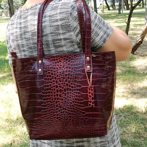 May include: A deep red tote bag with a glossy crocodile skin texture and pattern. The bag features two matching shoulder straps attached with gold-toned metal rivets and a small rectangular leather tag hanging from one of the strap attachments.