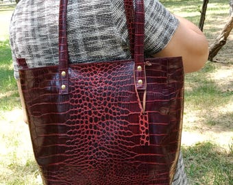 May include: A deep red tote bag with a glossy crocodile skin texture and pattern. The bag features two matching shoulder straps attached with gold-toned metal rivets and a small rectangular leather tag hanging from one of the strap attachments.
