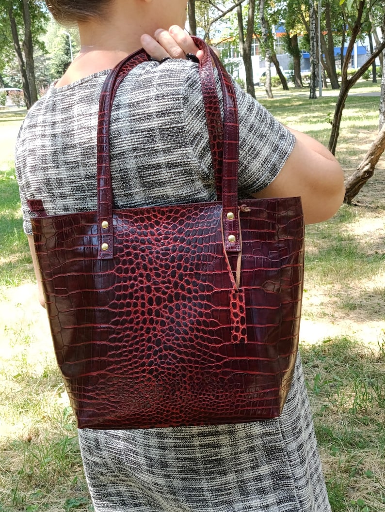 May include: A deep red tote bag with a glossy crocodile skin texture and pattern. The bag features two matching shoulder straps attached with gold-toned metal rivets and a small rectangular leather tag hanging from one of the strap attachments.