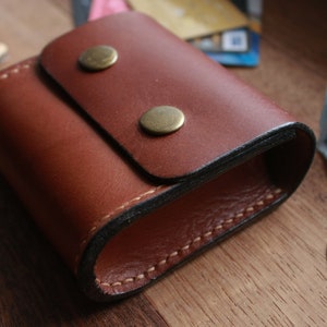 May include: A brown leather coin purse with a flap closure and two brass snaps. The purse is stitched with a black thread and has a dark brown lining.