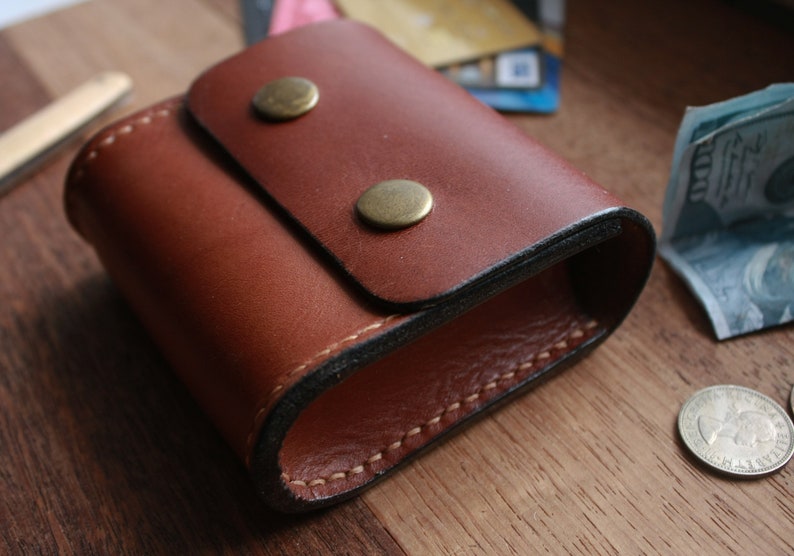 May include: A brown leather coin purse with a flap closure and two brass snaps. The purse is stitched with a black thread and has a dark brown lining.