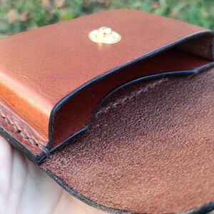 May include: A brown leather coin pouch featuring a smooth exterior finish and a textured interior surface. The pouch is secured with a circular gold-toned metal snap button fastener and displays visible dark stitching along the edges.