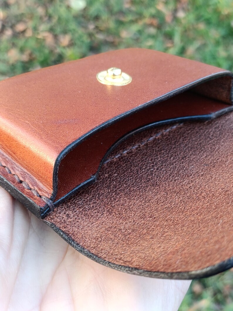 May include: A brown leather coin pouch featuring a smooth exterior finish and a textured interior surface. The pouch is secured with a circular gold-toned metal snap button fastener and displays visible dark stitching along the edges.