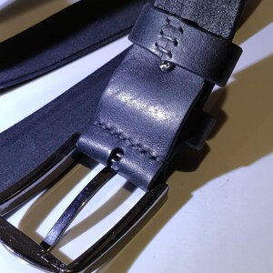May include: Close-up of a black leather belt with a silver-coloured buckle. The belt has a stitched loop and a metal pin. The leather appears to be textured. The buckle is rectangular with rounded corners.
