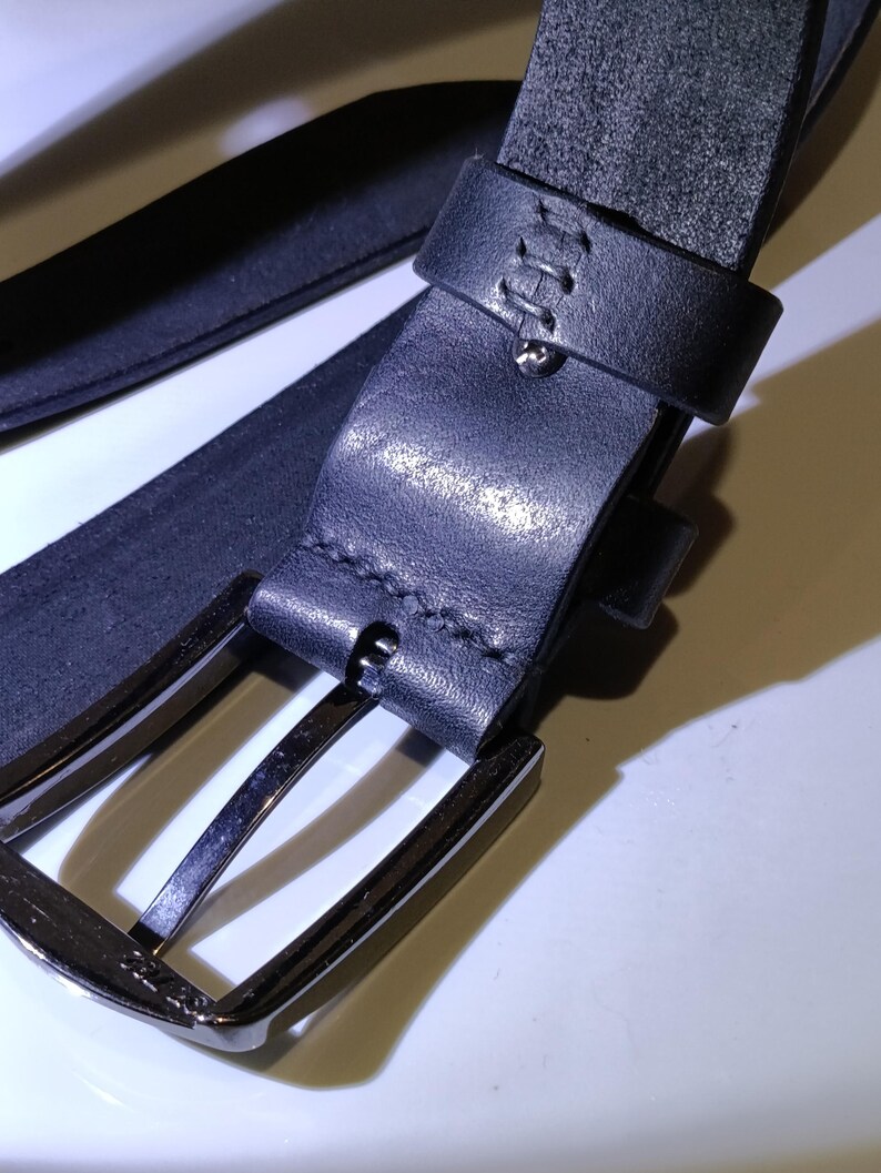 May include: Close-up of a black leather belt with a silver-coloured buckle. The belt has a stitched loop and a metal pin. The leather appears to be textured. The buckle is rectangular with rounded corners.