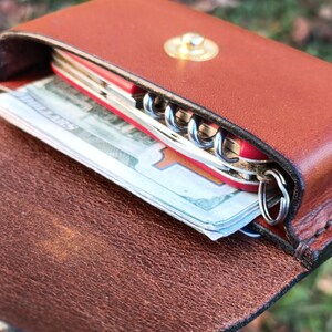 May include: A brown leather pouch with a gold-coloured snap closure containing a red multi-tool with a visible metal corkscrew and a folded stack of paper currency tucked inside the main compartment.