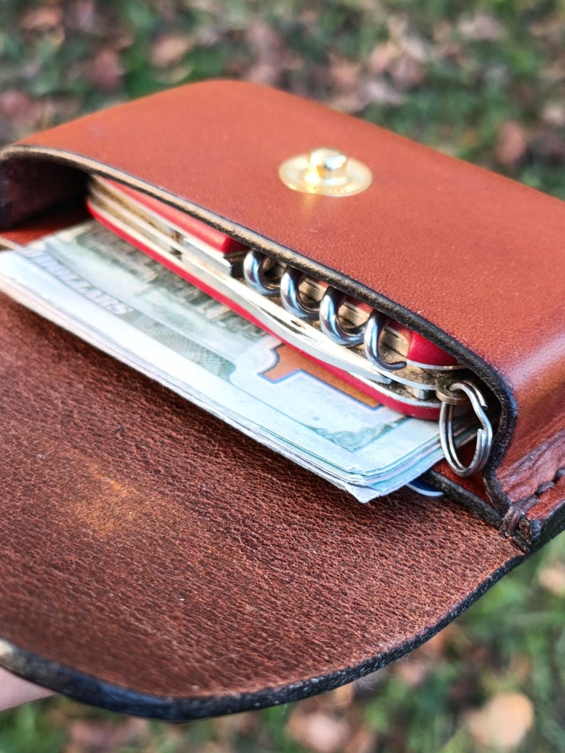 May include: A brown leather pouch with a gold-coloured snap closure containing a red multi-tool with a visible metal corkscrew and a folded stack of paper currency tucked inside the main compartment.