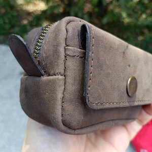 Peut inclure: Une petite pochette en cuir marron au fini mat, dot&eacute;e d'une fermeture &eacute;clair en m&eacute;tal couleur laiton avec une tirette en cuir, et d'une poche frontale &agrave; rabat ferm&eacute;e par un bouton-pression en m&eacute;tal couleur laiton. Coutures visibles sur les bords.