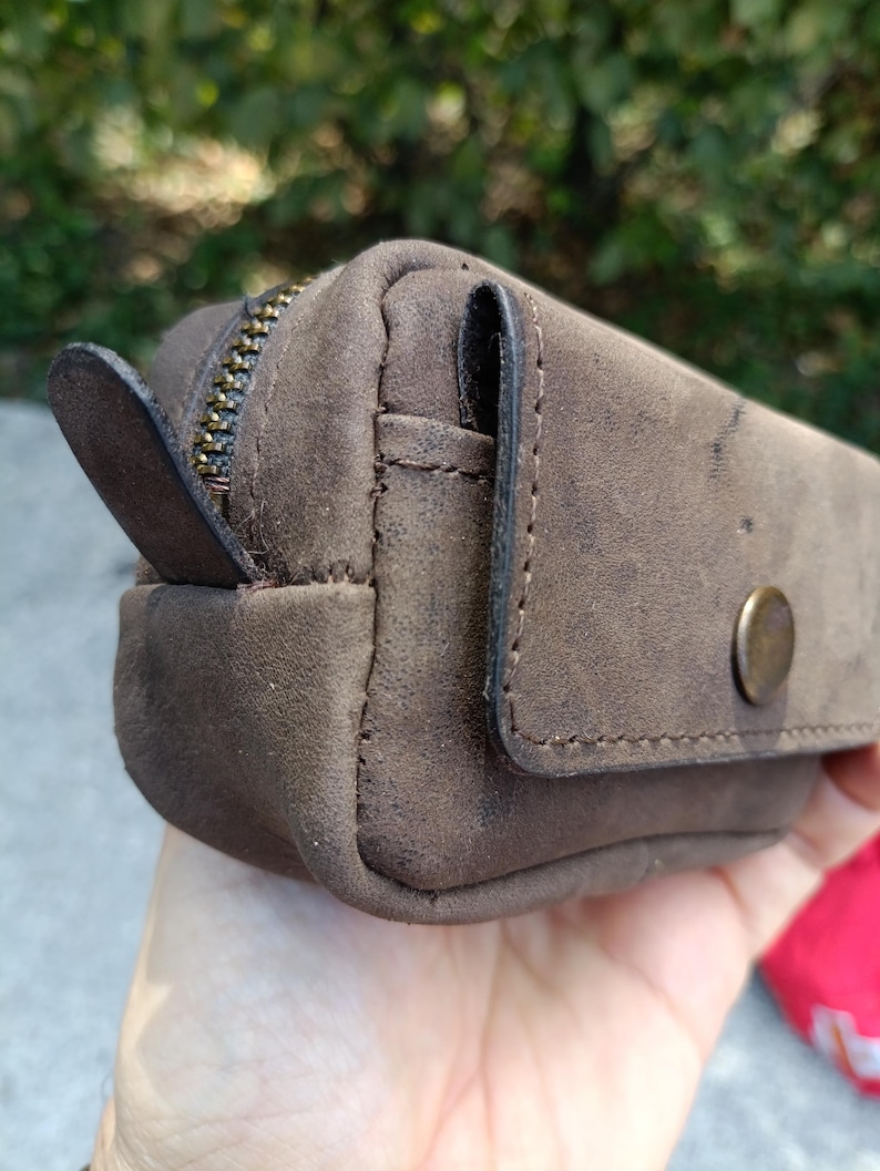 Peut inclure: Une petite pochette en cuir marron au fini mat, dot&eacute;e d'une fermeture &eacute;clair en m&eacute;tal couleur laiton avec une tirette en cuir, et d'une poche frontale &agrave; rabat ferm&eacute;e par un bouton-pression en m&eacute;tal couleur laiton. Coutures visibles sur les bords.