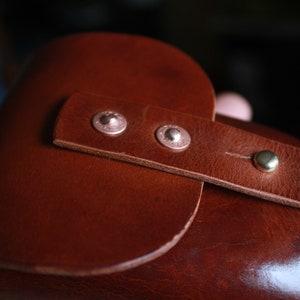 May include: Close-up of a brown leather item with a strap secured by two silver rivets and a brass button. The leather has a smooth texture and a rich, warm color, suggesting a handcrafted product.