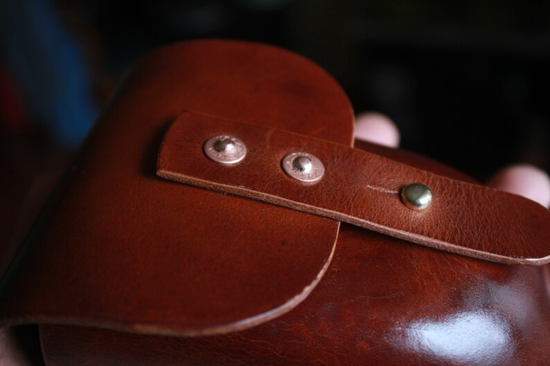 May include: Close-up of a brown leather item with a strap secured by two silver rivets and a brass button. The leather has a smooth texture and a rich, warm color, suggesting a handcrafted product.