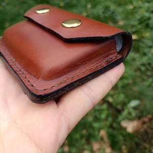 Puede incluir: Una peque&ntilde;a bolsa rectangular de cuero marr&oacute;n con cierre de solapa asegurado por dos botones de presi&oacute;n de lat&oacute;n pulido. Los bordes de la bolsa est&aacute;n acabados con costuras oscuras y bru&ntilde;idas, creando un dise&ntilde;o estructurado.