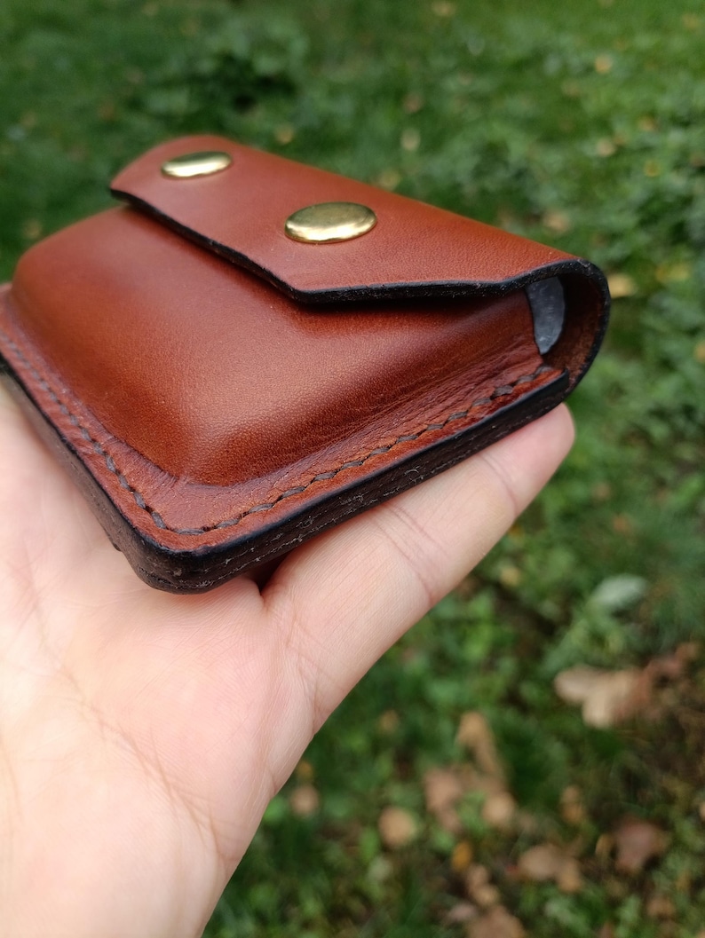 Puede incluir: Una peque&ntilde;a bolsa rectangular de cuero marr&oacute;n con cierre de solapa asegurado por dos botones de presi&oacute;n de lat&oacute;n pulido. Los bordes de la bolsa est&aacute;n acabados con costuras oscuras y bru&ntilde;idas, creando un dise&ntilde;o estructurado.