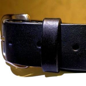 Op de afbeelding: Close-up van een zwarte leren riem met een zilverkleurige gesp. De riem heeft een klassiek ontwerp met een rechthoekige gesp en een lus om het uiteinde van de riem vast te zetten. Het leer lijkt glad en duurzaam.