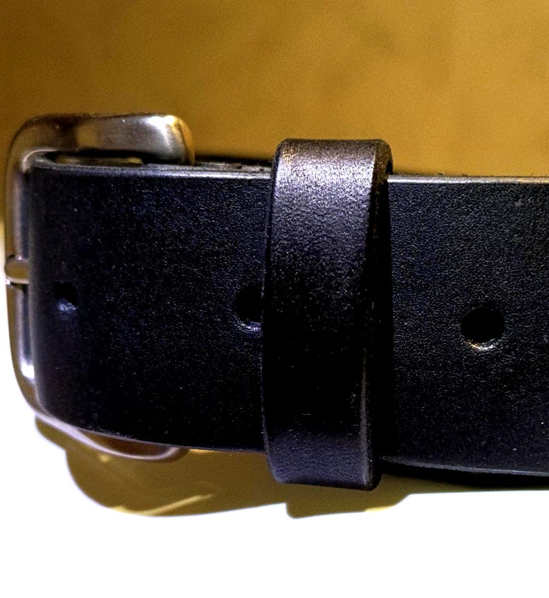 Op de afbeelding: Close-up van een zwarte leren riem met een zilverkleurige gesp. De riem heeft een klassiek ontwerp met een rechthoekige gesp en een lus om het uiteinde van de riem vast te zetten. Het leer lijkt glad en duurzaam.