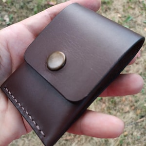 May include: A dark brown leather pouch featuring a smooth flap closure secured with a single round brass-toned metal snap button. The edges of the pouch are joined with visible, thick, light-colored stitching along the side and bottom seams.