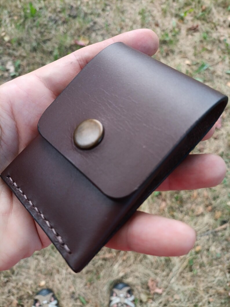 May include: A dark brown leather pouch featuring a smooth flap closure secured with a single round brass-toned metal snap button. The edges of the pouch are joined with visible, thick, light-colored stitching along the side and bottom seams.