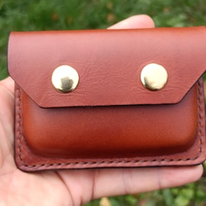 Puede incluir: Un estuche rectangular de cuero marr&oacute;n con cierre de solapa asegurado por dos botones de presi&oacute;n de metal dorado brillante. Los bordes del estuche presentan costuras oscuras visibles, lo que le otorga un dise&ntilde;o estructurado.