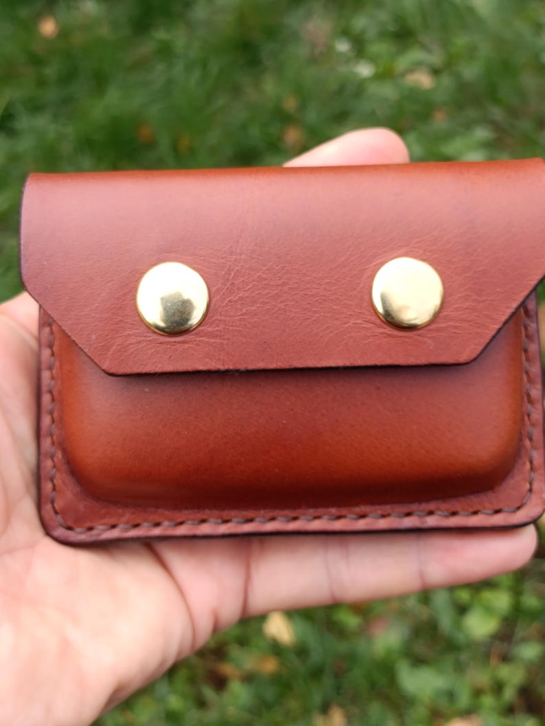Puede incluir: Un estuche rectangular de cuero marr&oacute;n con cierre de solapa asegurado por dos botones de presi&oacute;n de metal dorado brillante. Los bordes del estuche presentan costuras oscuras visibles, lo que le otorga un dise&ntilde;o estructurado.