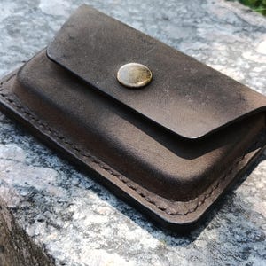 May include: A dark brown leather card holder with a rectangular shape and rounded corners. The accessory features visible stitching along the edges and a single metallic snap button closure centered on the front flap for secure fastening.