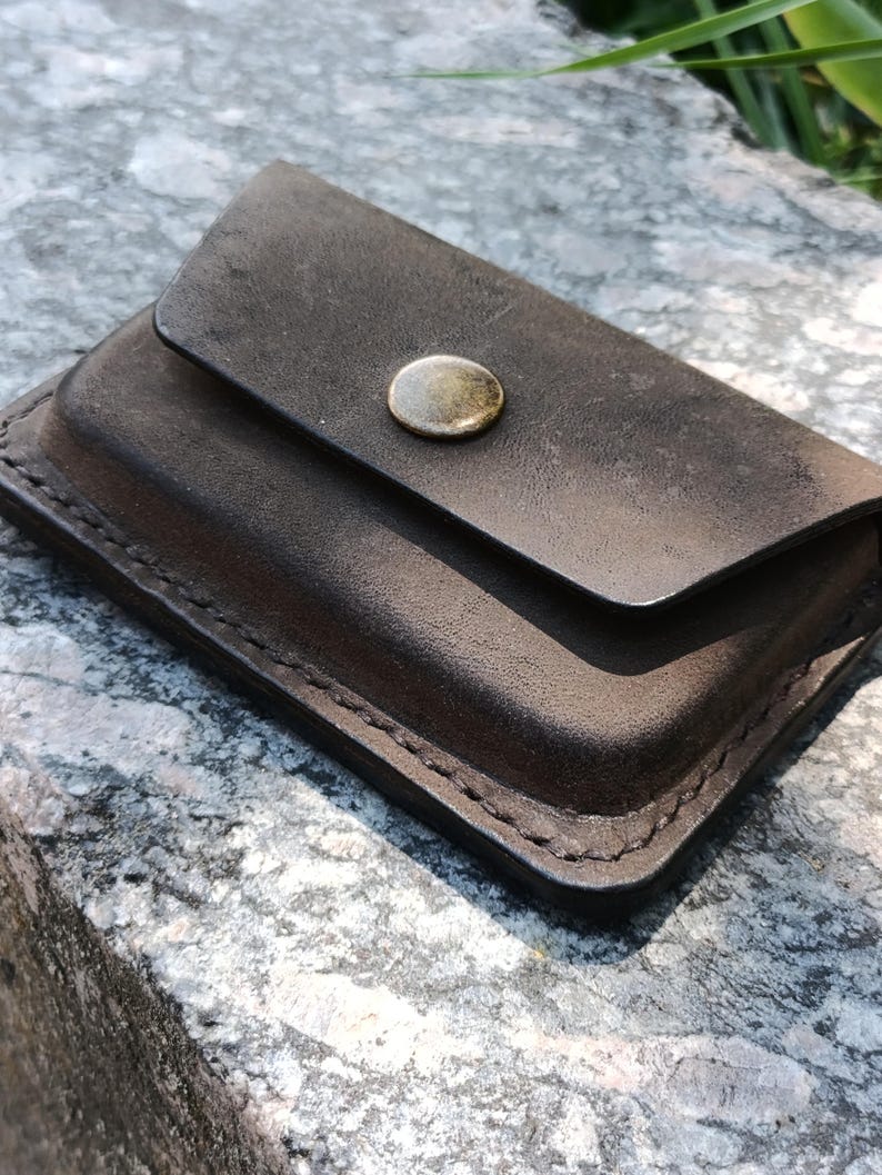May include: A dark brown leather card holder with a rectangular shape and rounded corners. The accessory features visible stitching along the edges and a single metallic snap button closure centered on the front flap for secure fastening.