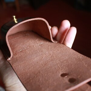 May include: A brown leather case, open to reveal the interior. The case has a curved top and a rectangular base. A small, gold-colored stud is visible on the top. The leather has a textured appearance.