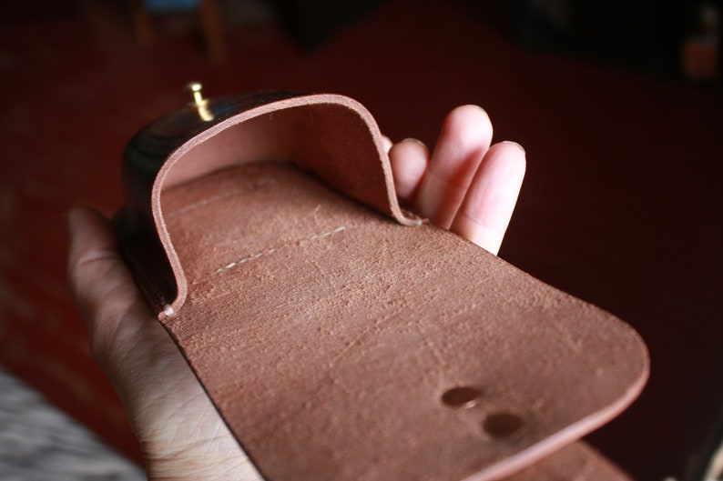 May include: A brown leather case, open to reveal the interior. The case has a curved top and a rectangular base. A small, gold-colored stud is visible on the top. The leather has a textured appearance.
