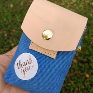 May include: A blue canvas pouch featuring a light tan leather flap secured with a gold-tone metal snap button. A circular white sticker is attached to the front with the text thank you printed in a metallic copper script font.