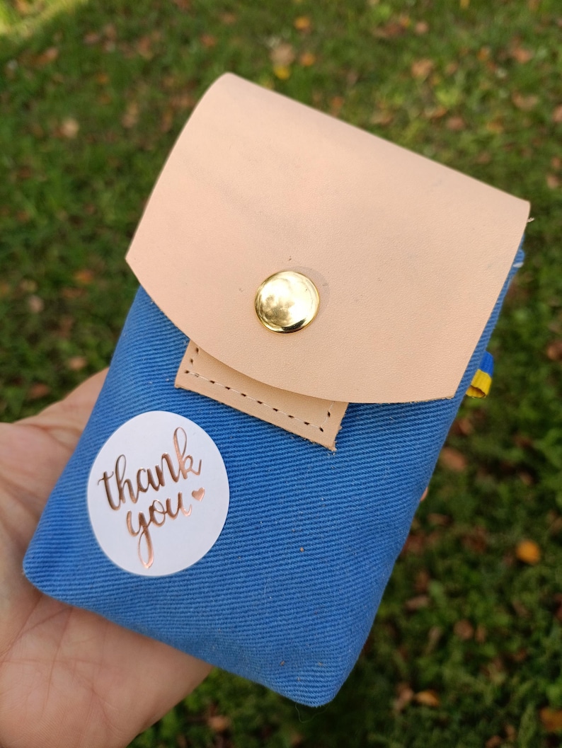 May include: A blue canvas pouch featuring a light tan leather flap secured with a gold-tone metal snap button. A circular white sticker is attached to the front with the text thank you printed in a metallic copper script font.
