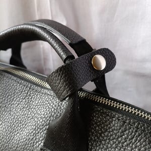 Puede incluir: Primer plano de un bolso de cuero negro con una superficie texturizada. El bolso presenta una cremallera plateada y un asa con una correa negra asegurada por un bot&oacute;n plateado. El bolso est&aacute; dise&ntilde;ado para transportar objetos.