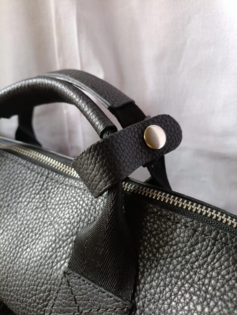 Puede incluir: Primer plano de un bolso de cuero negro con una superficie texturizada. El bolso presenta una cremallera plateada y un asa con una correa negra asegurada por un bot&oacute;n plateado. El bolso est&aacute; dise&ntilde;ado para transportar objetos.