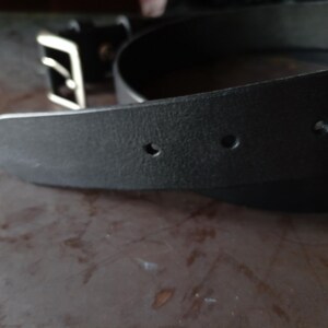 Puede incluir: Un cintur&oacute;n de cuero negro con superficie texturizada y punta afilada que presenta tres orificios de ajuste circulares visibles. El cintur&oacute;n incluye una hebilla rectangular de metal plateado con un solo perno en el extremo.