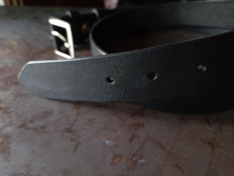 Puede incluir: Un cintur&oacute;n de cuero negro con superficie texturizada y punta afilada que presenta tres orificios de ajuste circulares visibles. El cintur&oacute;n incluye una hebilla rectangular de metal plateado con un solo perno en el extremo.
